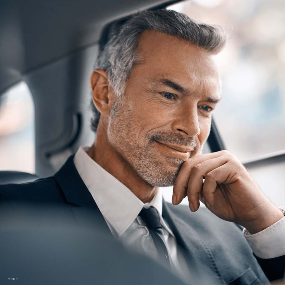 Middle aged man with peppered grey hair in a tie and suit sitting in a car