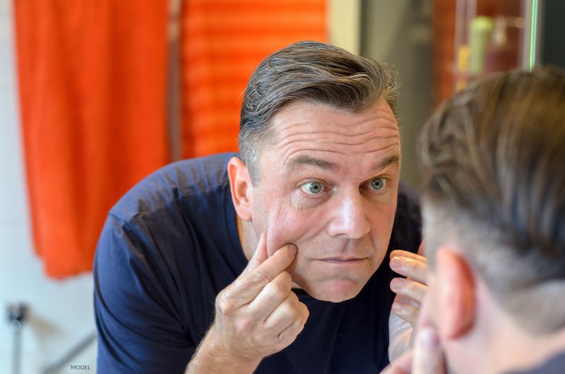 Middle-aged man looks at his reflection in the mirror while he pinches sagging skin in his cheeks.